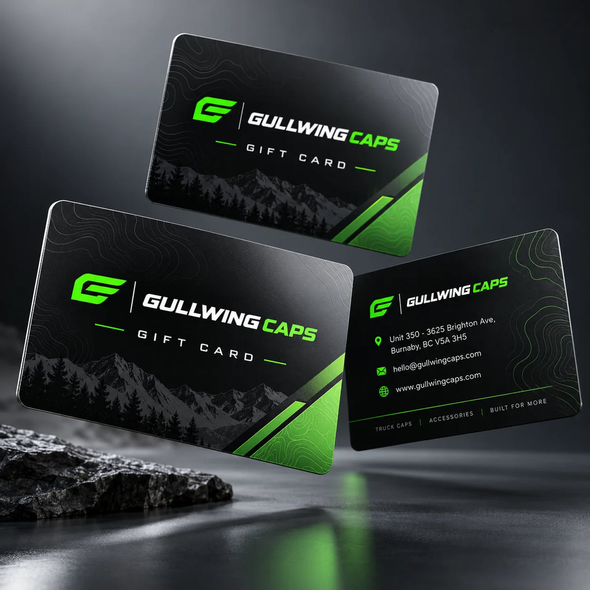 Three Gullwing Caps gift cards are displayed in a floating arrangement on a dark reflective surface with a rock prop. The cards feature a black and dark gray mountain pattern with bright green accents and white text.