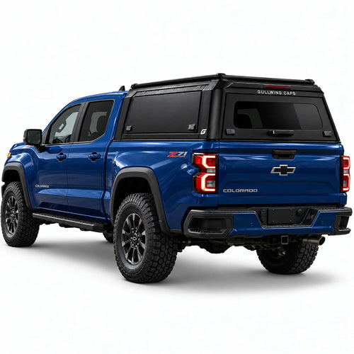 A blue Chevrolet Colorado pickup truck shown from a rear three-quarter angle, fitted with a black Gullwing Caps aerowing truck canopy, set against a plain white studio background.