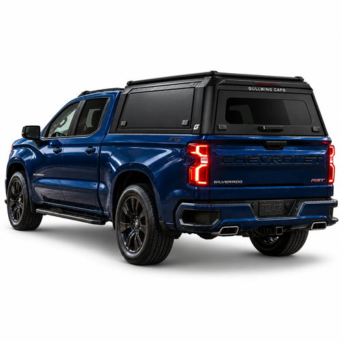 A blue Chevrolet Silverado 1500 pickup truck shown from a rear three-quarter angle, fitted with a black Gullwing Caps aerowing truck canopy, set against a plain white studio background.