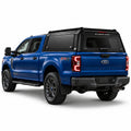 A blue Ford f250 Super Duty FX4 pickup truck shown from a rear three-quarter angle, fitted with a black Gullwing Caps aerowing truck canopy, set against a plain white studio background.