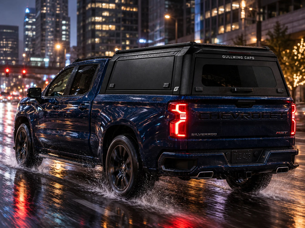 A dark blue Chevrolet Silverado pickup truck with a black Gullwing Caps canopy, shown from a rear three-quarter angle while driving through splashing rain on a wet city street at night.