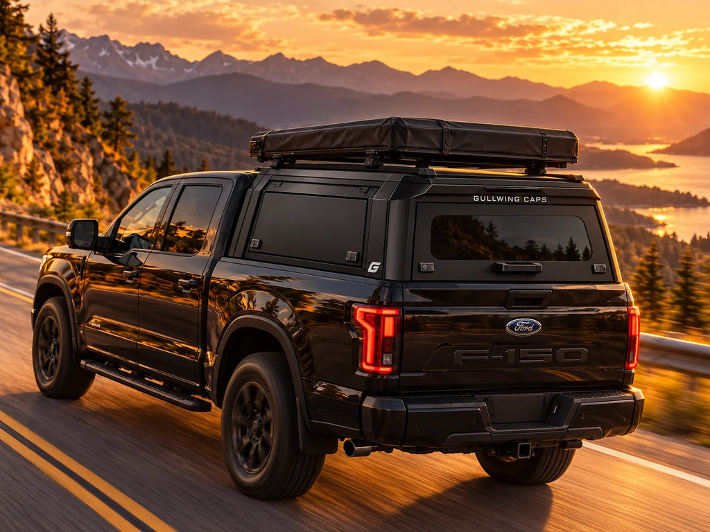 A black Ford F-150 pickup truck with a roof-mounted black tent is driving on a mountain road at sunset. The rear window has 'GULLWING CAPS' branding, and the tailgate shows the Ford F-150 badge.