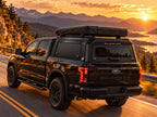 A black Ford F-150 pickup truck with a roof-mounted black tent is driving on a mountain road at sunset. The rear window has 'GULLWING CAPS' branding, and the tailgate shows the Ford F-150 badge.