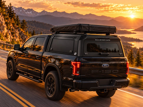 A black Ford F-150 pickup truck with a roof-mounted black tent is driving on a mountain road at sunset. The rear window has 'GULLWING CAPS' branding, and the tailgate shows the Ford F-150 badge.