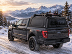 A black Ford F-150 pickup truck with a black gullwing Caps canopy over the bed, parked on a snowy mountain road with snow-covered peaks in the background.