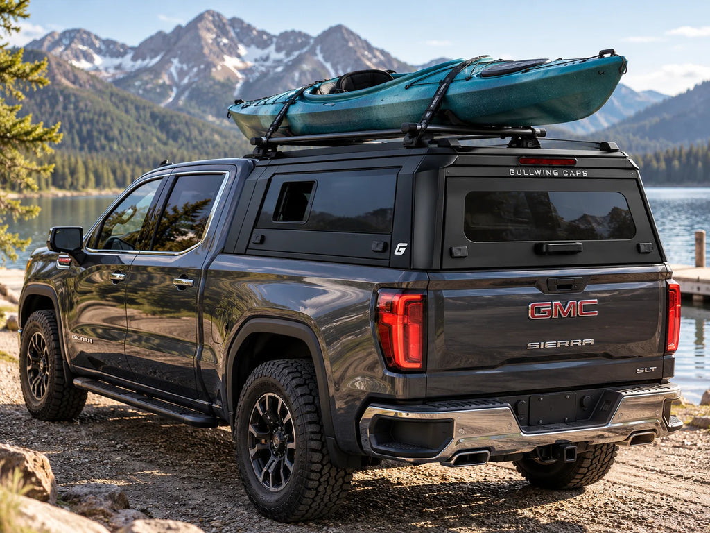 A dark gray GMC Sierra pickup truck parked outdoors beside a lake with mountains in the background with a gullwing caps sliding window truck canopy and kayak strapped to the top. The truck is shown from a rear three-quarter angle, equipped with a black roof rack carrying a teal kayak.