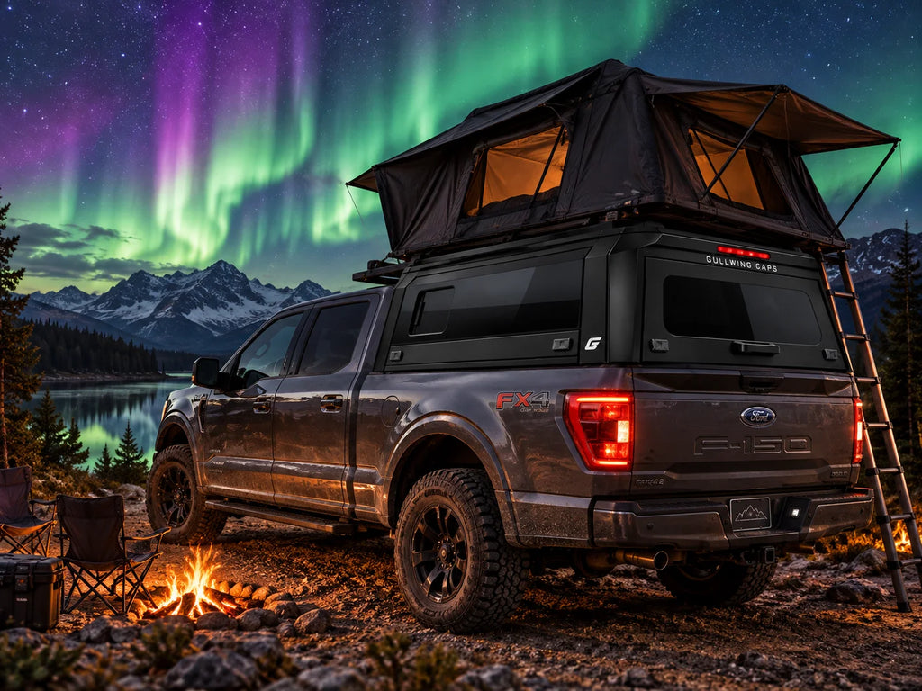 A dark gray Ford F-150 pickup truck with a rooftop tent and gullwing caps sliding window canopy is parked outdoors at night beside a campfire, with mountains, water, and aurora lights in the sky in the background.
