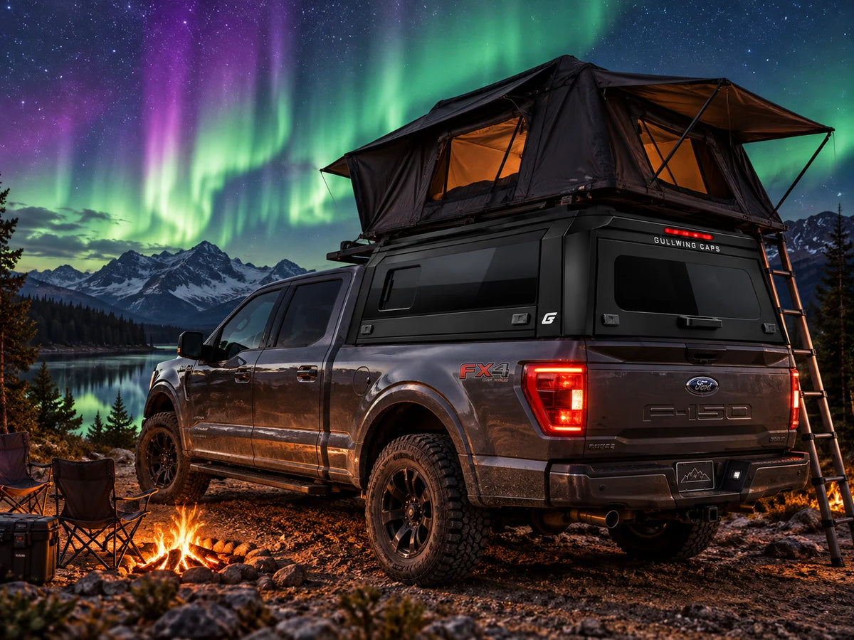 A dark gray Ford F-150 pickup truck with a rooftop tent and gullwing caps sliding window canopy is parked outdoors at night beside a campfire, with mountains, water, and aurora lights in the sky in the background.