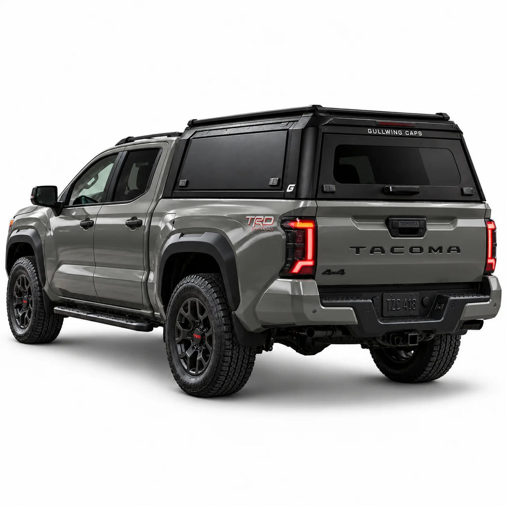 A gray Toyota Tacoma pickup truck shown from a rear three-quarter angle, fitted with a black Gullwing Caps aerowing truck canopy, set against a plain white studio background.