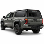 A gray Toyota Tacoma pickup truck shown from a rear three-quarter angle, fitted with a black Gullwing Caps aerowing truck canopy, set against a plain white studio background.