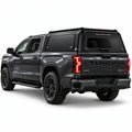 A dark gray Tundra 4x4 pickup truck shown from a rear three-quarter angle, fitted with a black Gullwing Caps aerowing truck canopy. The truck features black wheels, black side steps, and red accents on the TRD badge and wheel centers, set against a plain white studio background.
