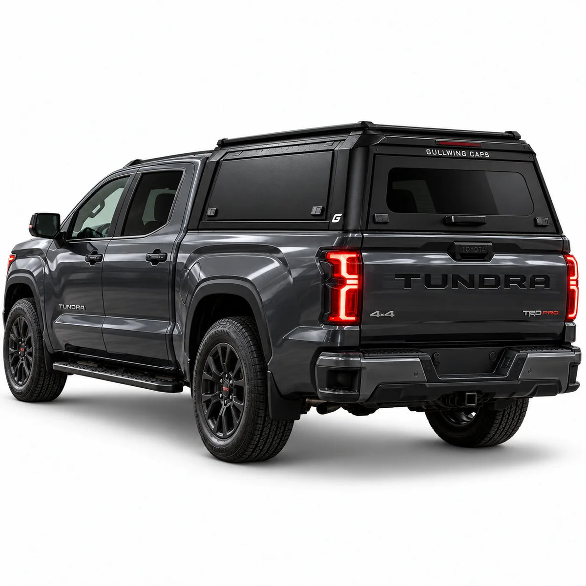 A dark gray Tundra 4x4 pickup truck shown from a rear three-quarter angle, fitted with a black Gullwing Caps aerowing truck canopy. The truck features black wheels, black side steps, and red accents on the TRD badge and wheel centers, set against a plain white studio background.